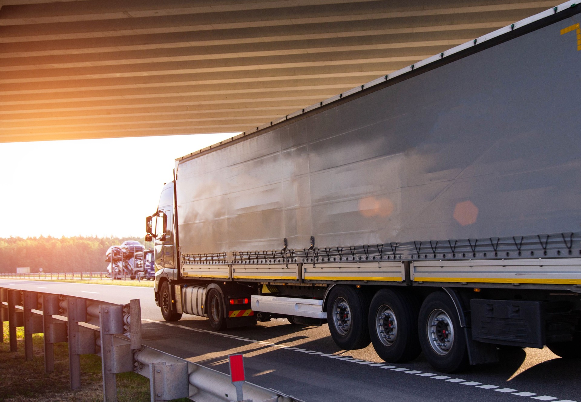 A truck with a tilt semi-trailer carries cargo under a bridge against the backdrop of sunset, copy space for text. Logistics